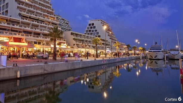 Un paseo marítimo con edificios modernos, un puerto deportivo con yates y una masa de agua tranquila que refleja las construcciones.