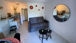 Un espacio habitable pequeño con un sofá gris, dos mesas redondas y una cocina visible a través de una puerta abierta.