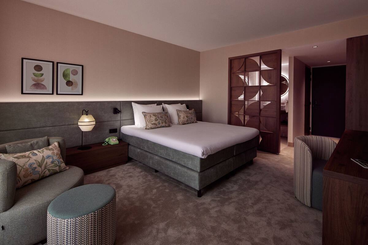 A modern bedroom with a large bed, a sofa, a small ottoman, and a wooden partition.