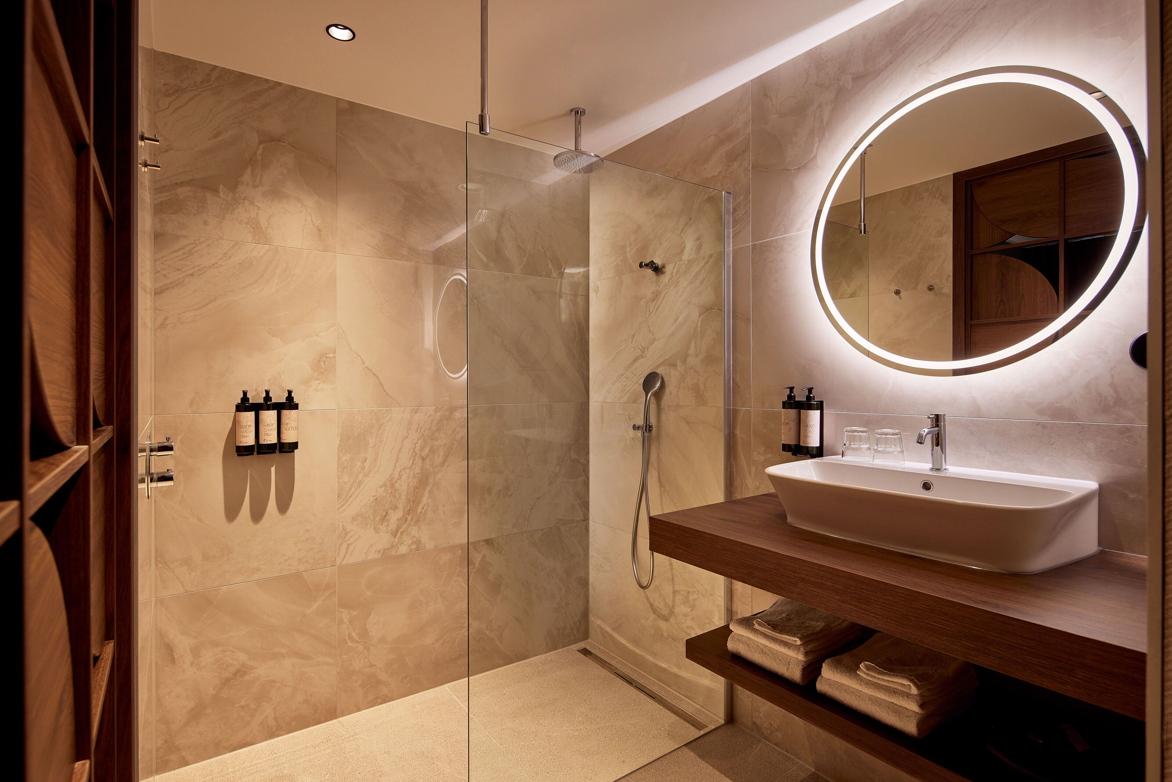 Un baño moderno con ducha envuelta en vidrio, tocador con lavamanos y espejo redondo con luz.