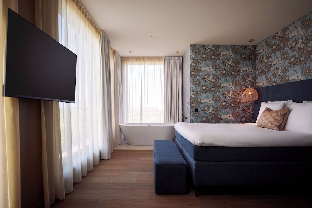 A bedroom with a large bed, a flat-screen TV, a bathtub, and a window with curtains.