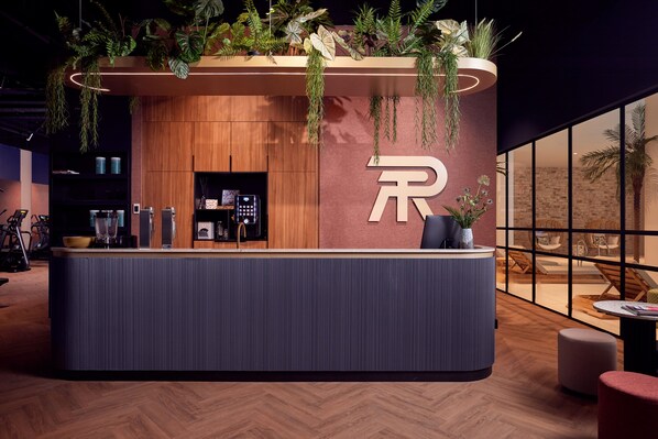 A reception desk with a wooden finish, a large 'R' logo, and hanging plants. - Van der Valk Hotel Lelystad (Lelystad)