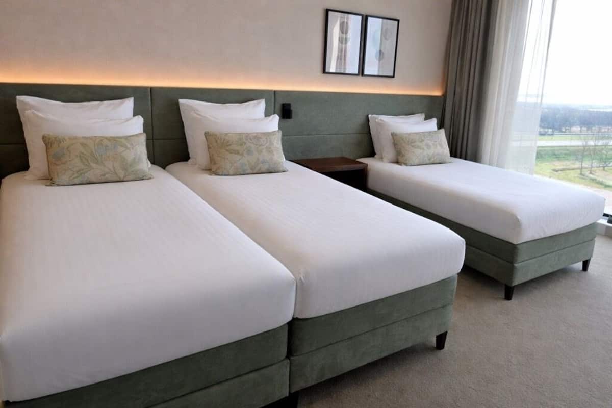A hotel room with two single beds, a nightstand, a wall-mounted light, and a window with a view.