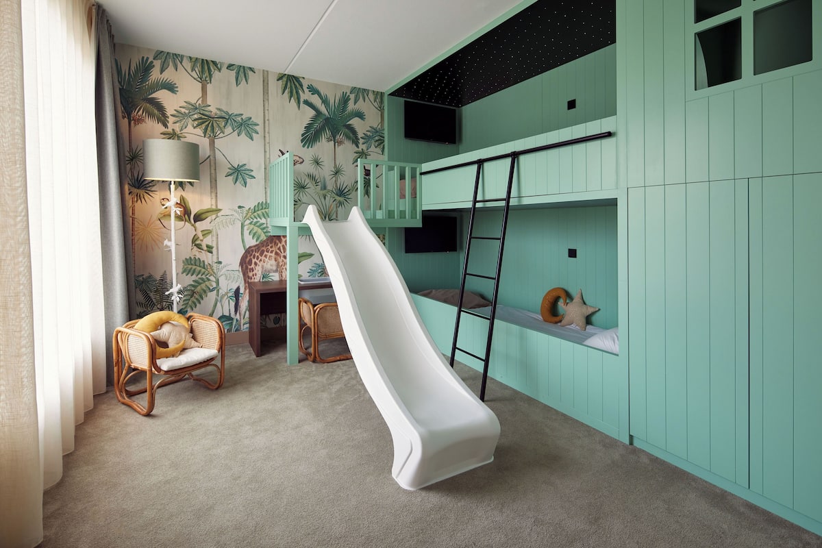 A child's playroom with a slide, bunk beds, a rattan chair, and a wall mural.