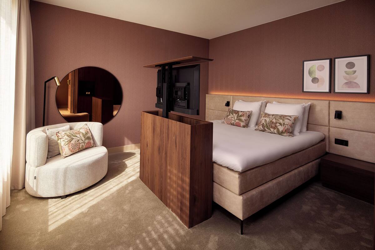 A modern hotel room with a curved sofa, a large bed with a headboard, a wooden TV stand, and a round mirror.