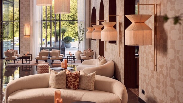 A modern lounge area with a large beige sofa, multiple armchairs, and small tables. There are decorative candles and a large window providing a view of the outdoors. - Van der Valk Hotel Lelystad (Lelystad)