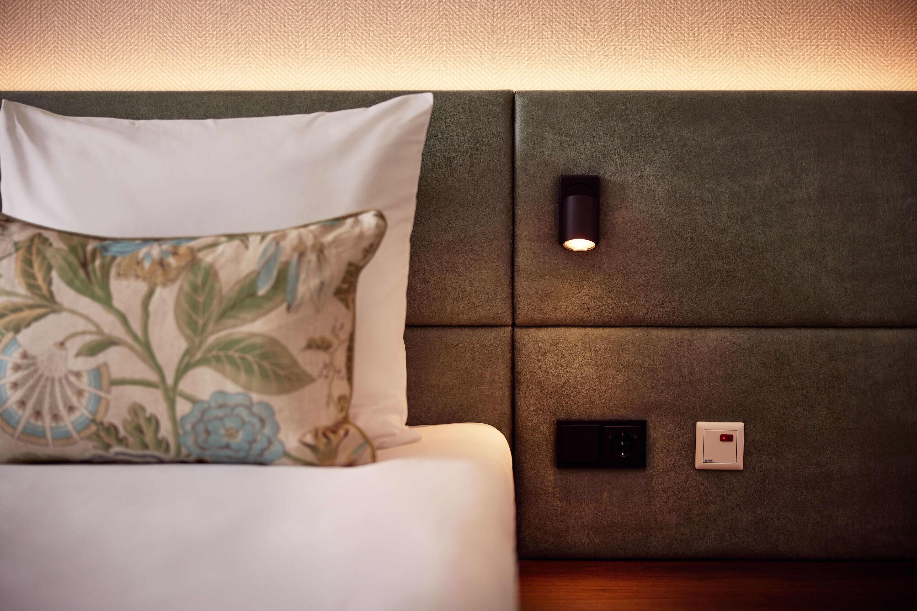 Una cama con un cojín estampado, una lámpara fijada a la pared y un termostato.