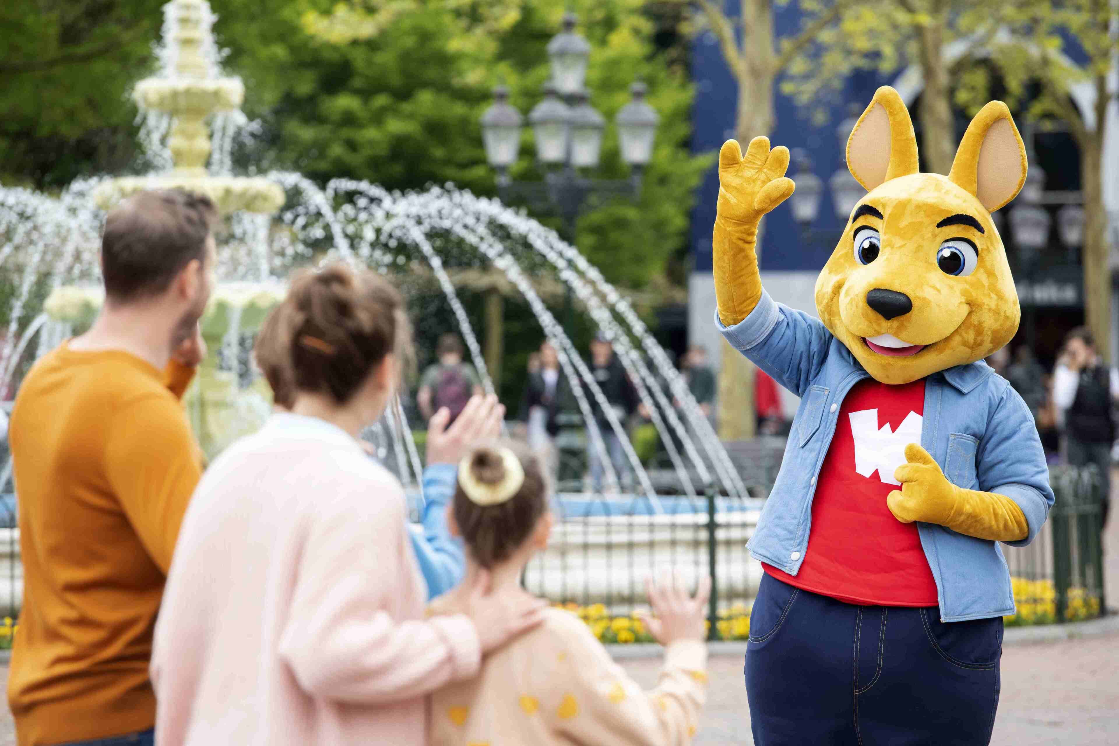 Une mascotte dans un parc avec une fontaine, et des gens en arrière-plan.