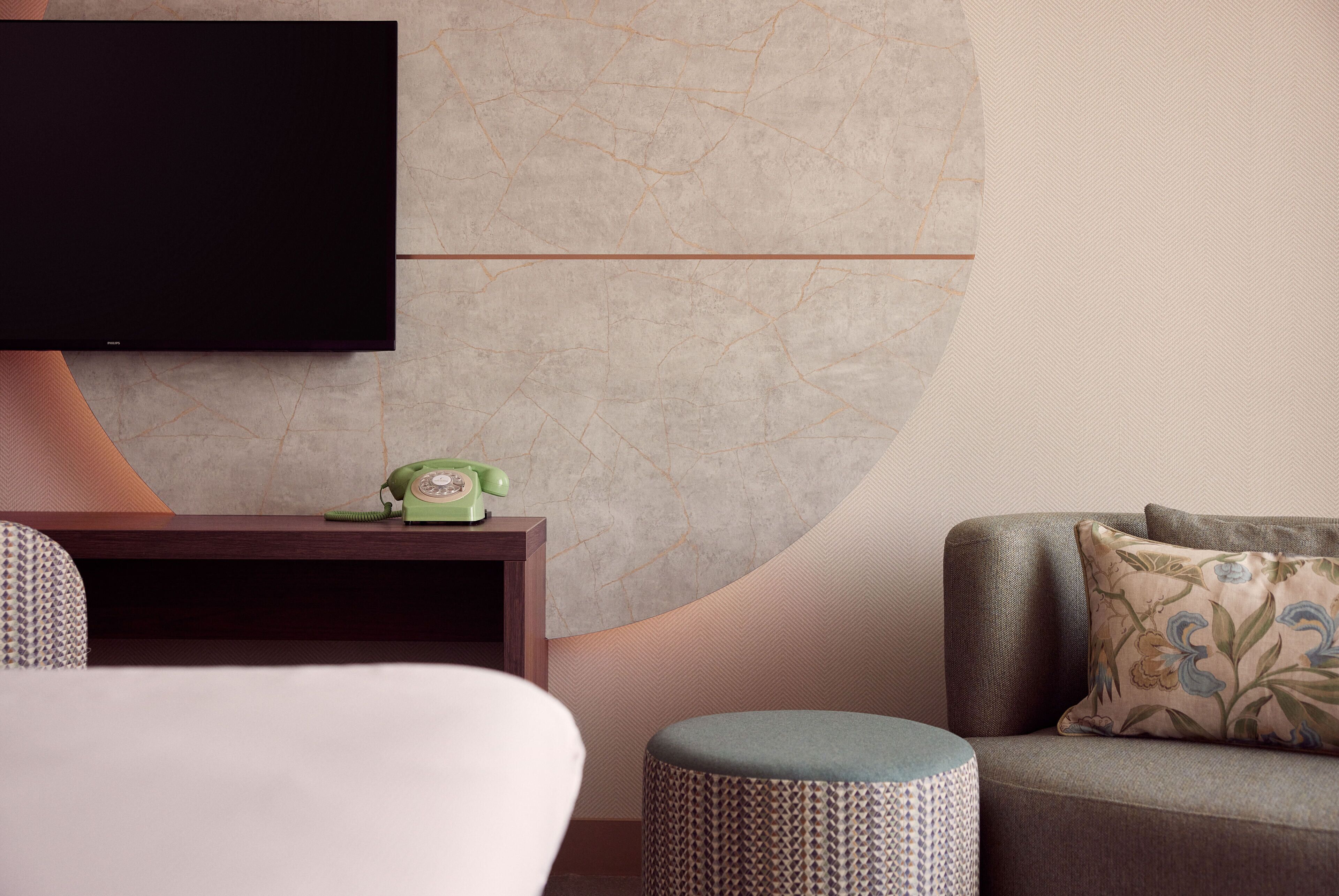 A modern hotel room with a flat-screen TV mounted on the wall, a wooden nightstand with a vintage telephone, a patterned ottoman, and a sofa with floral cushions.
