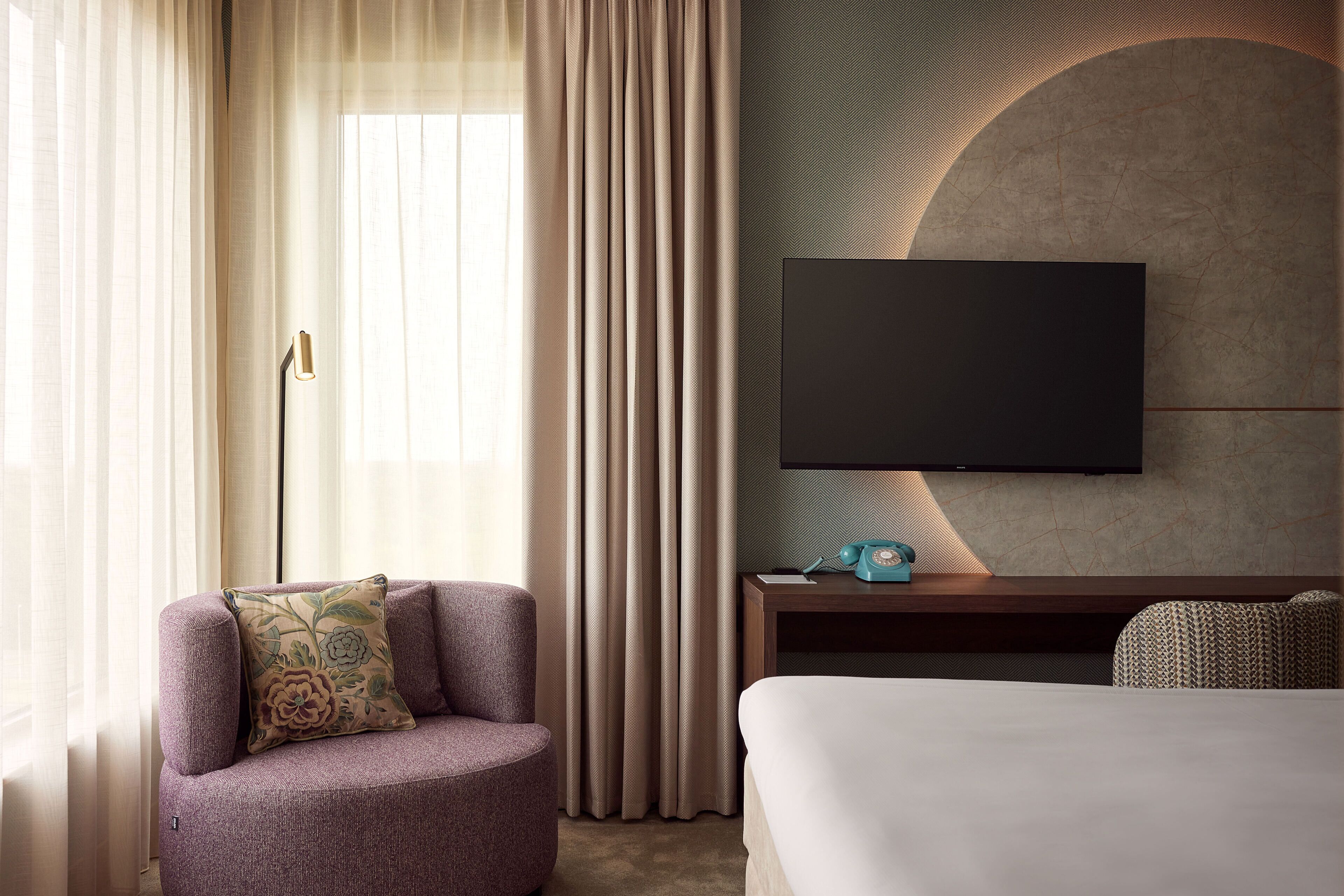 Ein modernes Hotelzimmer mit einem Bett, einer Couch, einem an der Wand montierten Fernseher und einem Nachttisch mit Lampe.