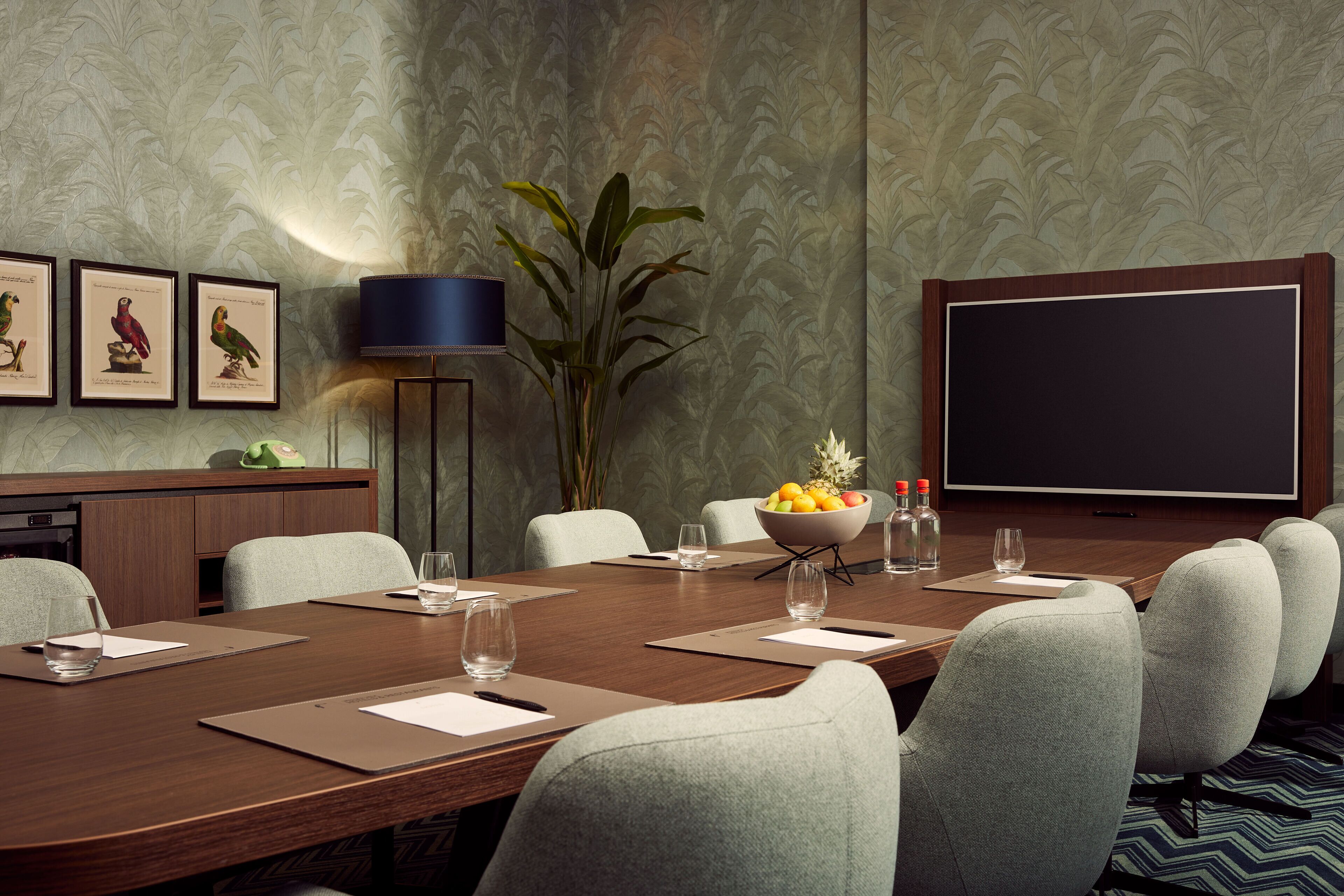 A conference room with a wooden table, grey upholstered chairs, a projector screen, framed bird illustrations, a plant, and a fruit bowl.