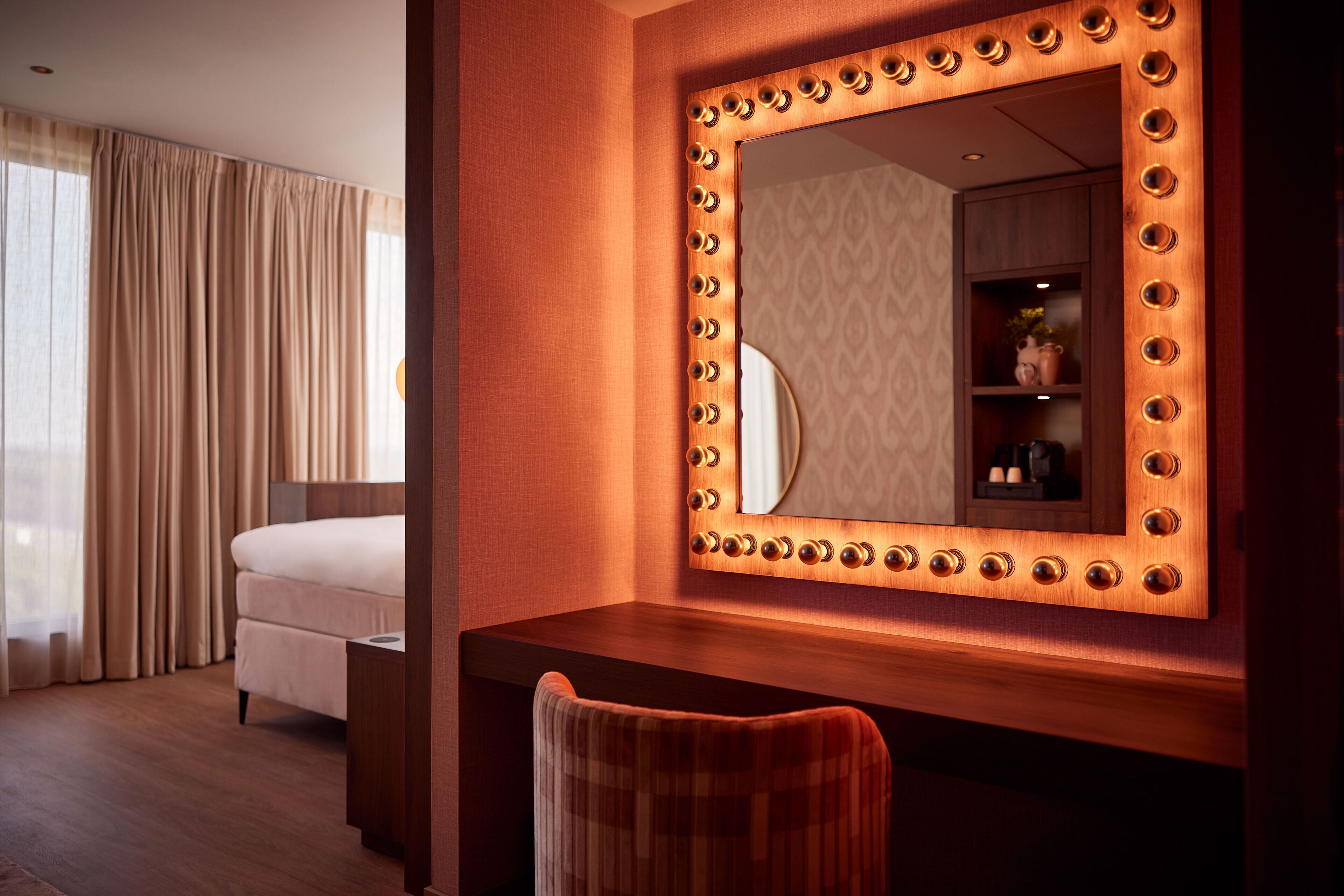 A hotel room with a vanity area, a bed, and a large mirror with lights.