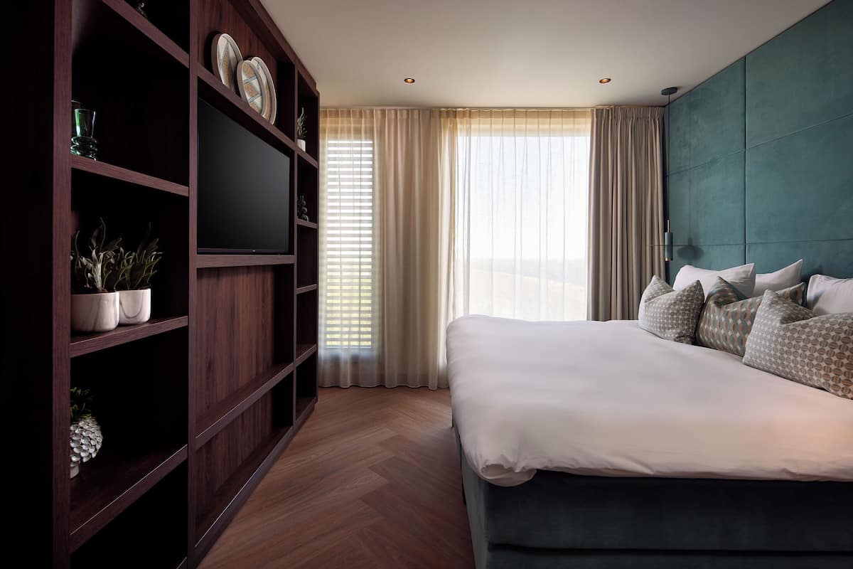 A modern hotel room with a large bed, a TV on a wooden shelf, and a view of the city through the window.