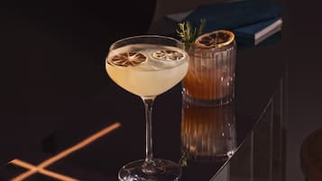 Deux verres contenant une boisson claire agrémentée de tranches d’agrumes séchées, un verre contenant un liquide ambré et une branche de romarin, le tout posé sur une surface réfléchissante.