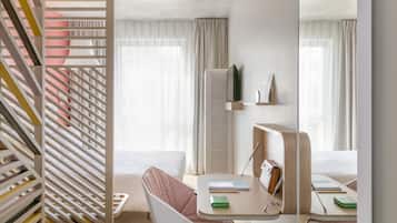 A modern hotel room with a bed, a desk with a chair, a mirror, and a window with curtains.