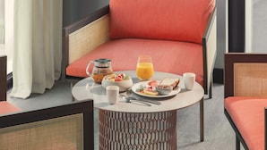 A breakfast setup with a wicker chair, a round table, and a breakfast tray including cereal, toast, fruit, and juice. - OKKO Hotels Paris la Défense (Nanterre)