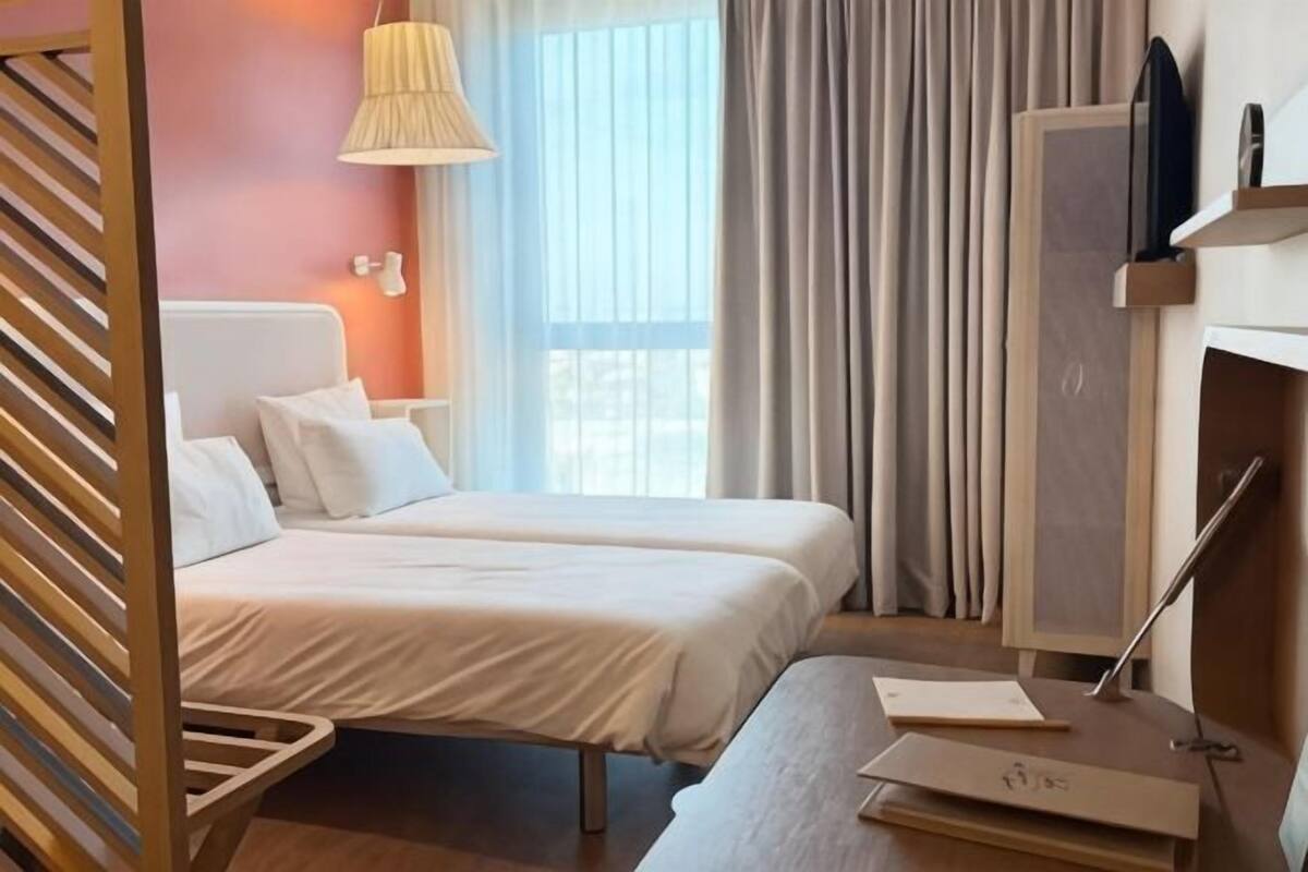 Habitación de hotel con dos camas, un escritorio, una lámpara, un televisor y una ventana con cortinas.