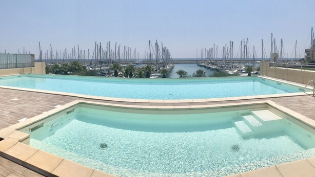 Una piscina en la azotea con agua cristalina, rodeada por una terraza de madera y vistas a un puerto deportivo con numerosos barcos.
