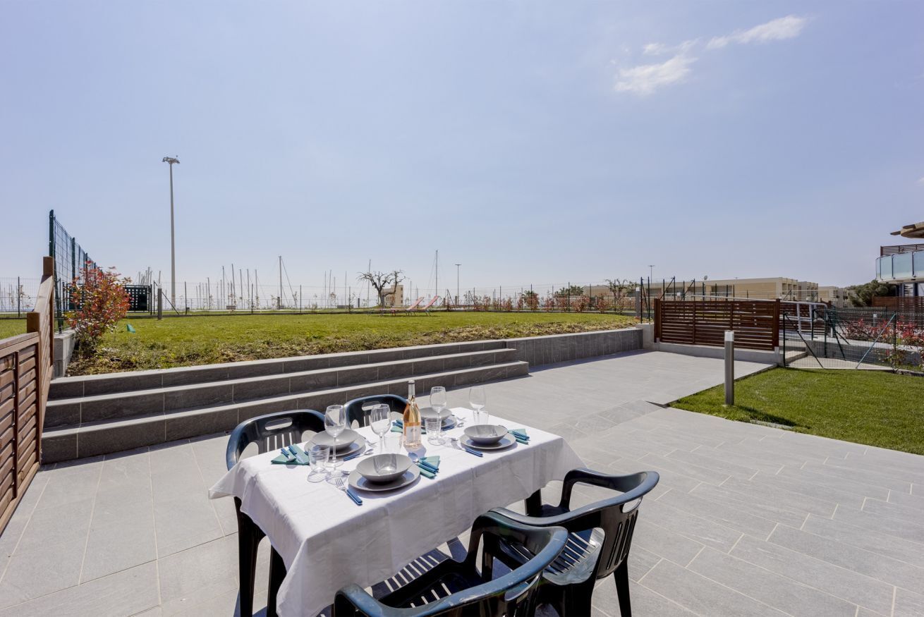 Un espace extérieur aménagé pour les repas, avec une table dressée pour quatre personnes, entourée de chaises, sur une terrasse offrant une vue sur un jardin paysager et des bâtiments situés au loin.