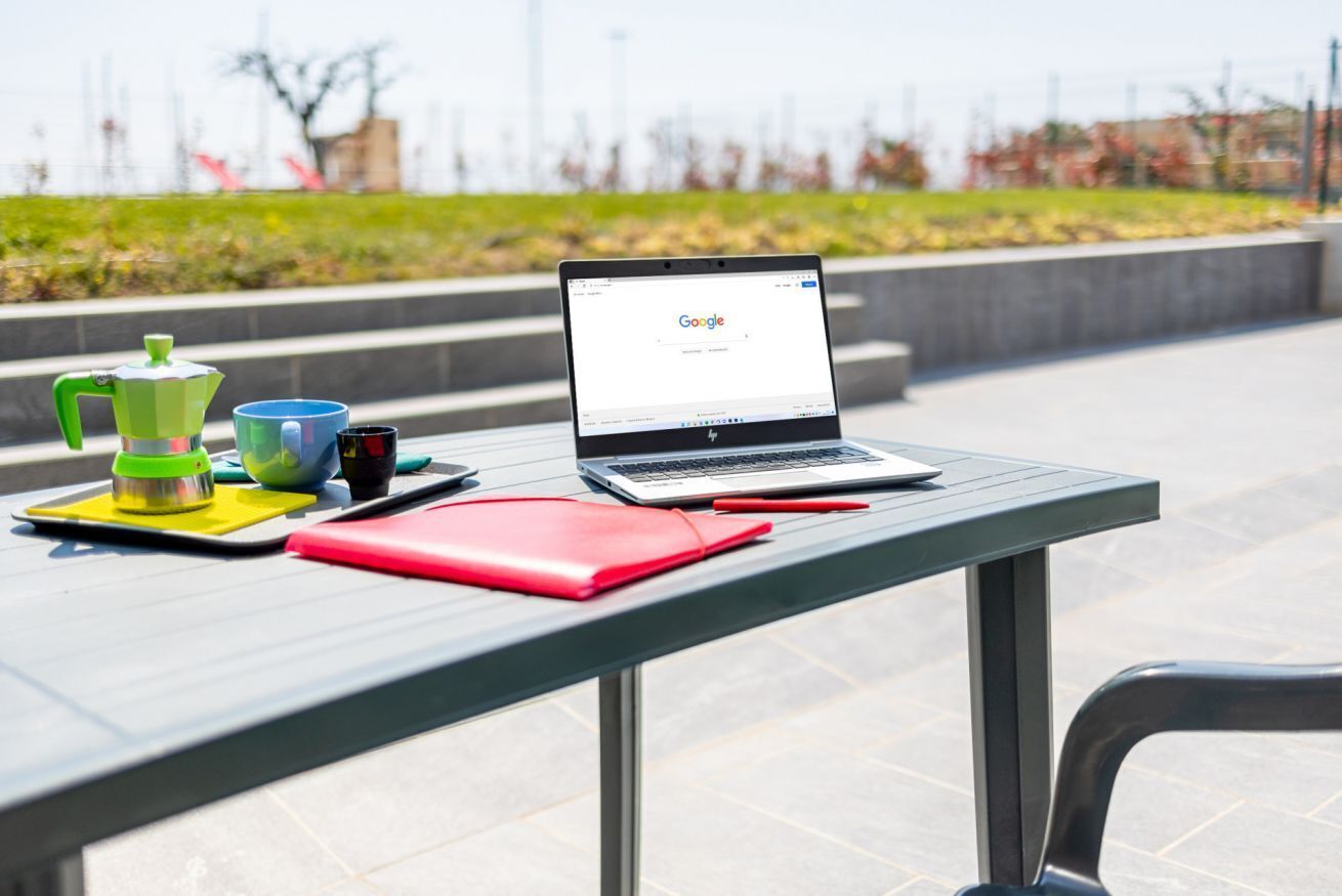 Una laptop sobre una mesa con una taza verde y azul, un cuaderno rojo y una taza negra.