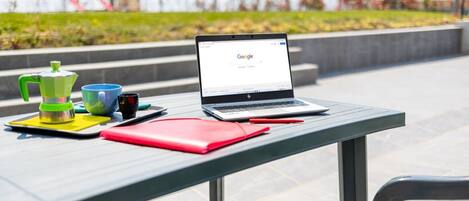Una laptop sobre una mesa con una taza verde y azul, un cuaderno rojo y una taza negra.