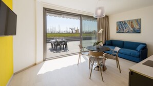 Ein modernes Wohnzimmer mit einem blauen Sofa, einem Esstisch mit Stühlen, einem großen Fenster mit Meerblick und einem Gemälde an der Wand.