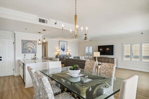 A spacious living area with a glass dining table, comfortable seating, a modern chandelier, and a flat-screen TV.