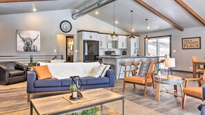 A modern living room with a sofa, coffee table, and chairs. There is a kitchen area with cabinets and a refrigerator. A clock and a sign on the wall are visible. - Modern Lead Cabin w/ Private Hot Tub & Fireplace! (Lead)