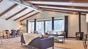 A spacious living room with a high ceiling, large windows, a fireplace, and a dining area with wooden furniture. - Modern Lead Cabin w/ Private Hot Tub & Fireplace! (Lead)