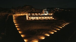 Un grand bâtiment illuminé, doté de nombreuses fenêtres et d’une tour centrale, entouré d’une zone pavée et d’un terrain rocheux.