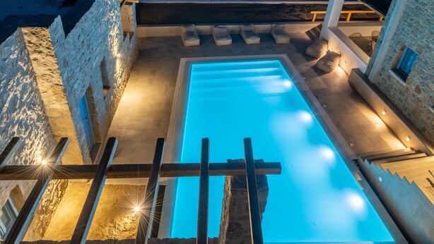 Une piscine avec des murs en pierre, une terrasse en bois et des sièges d’extérieur.