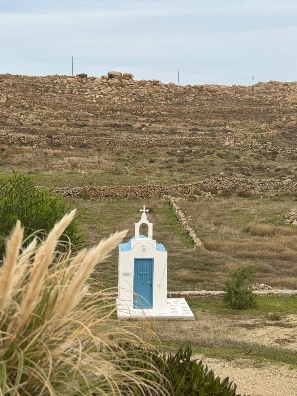 A small white church with a blue door and a cross on top, surrounded by dry grass and a rocky hill in the background.