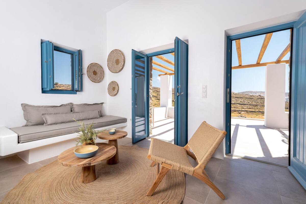 Una sala moderna con un sofá, una silla de fibras naturales, una mesa redonda de madera y vistas a una terraza exterior a través de puertas abiertas.
