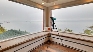 A room with large windows overlooking a body of water, a telescope on a tripod, and a cushioned bench with a pillow. - Chic Port Angeles Home w/ Oceanfront Balcony! (Port Angeles)