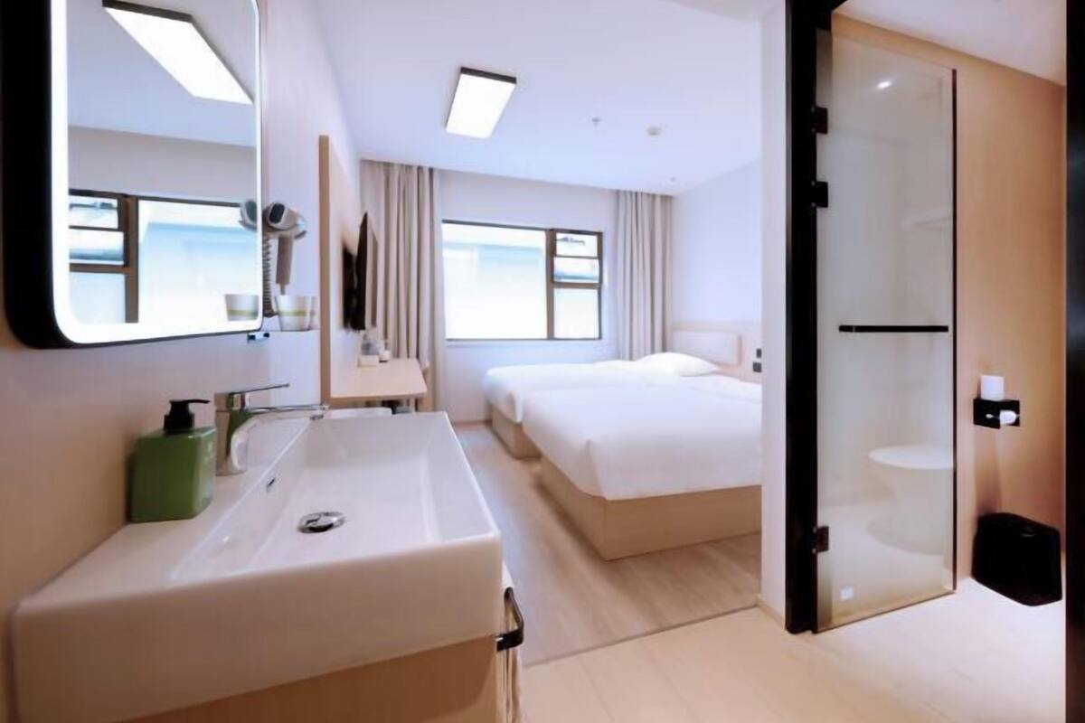 Habitación de hotel con un espejo grande, un lavabo, dos camas, una puerta de ducha de vidrio y vistas al exterior.