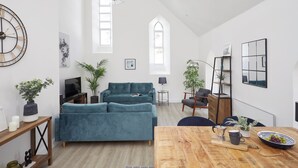 A spacious living room with a large teal sofa, wooden coffee table, and a TV. There are two windows with arched tops, a chandelier, and a bookshelf. - No 5 Old Church (Ventnor)