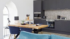 A modern kitchen with dark cabinets, a wooden dining table, and blue chairs. - No 5 Old Church (Ventnor)