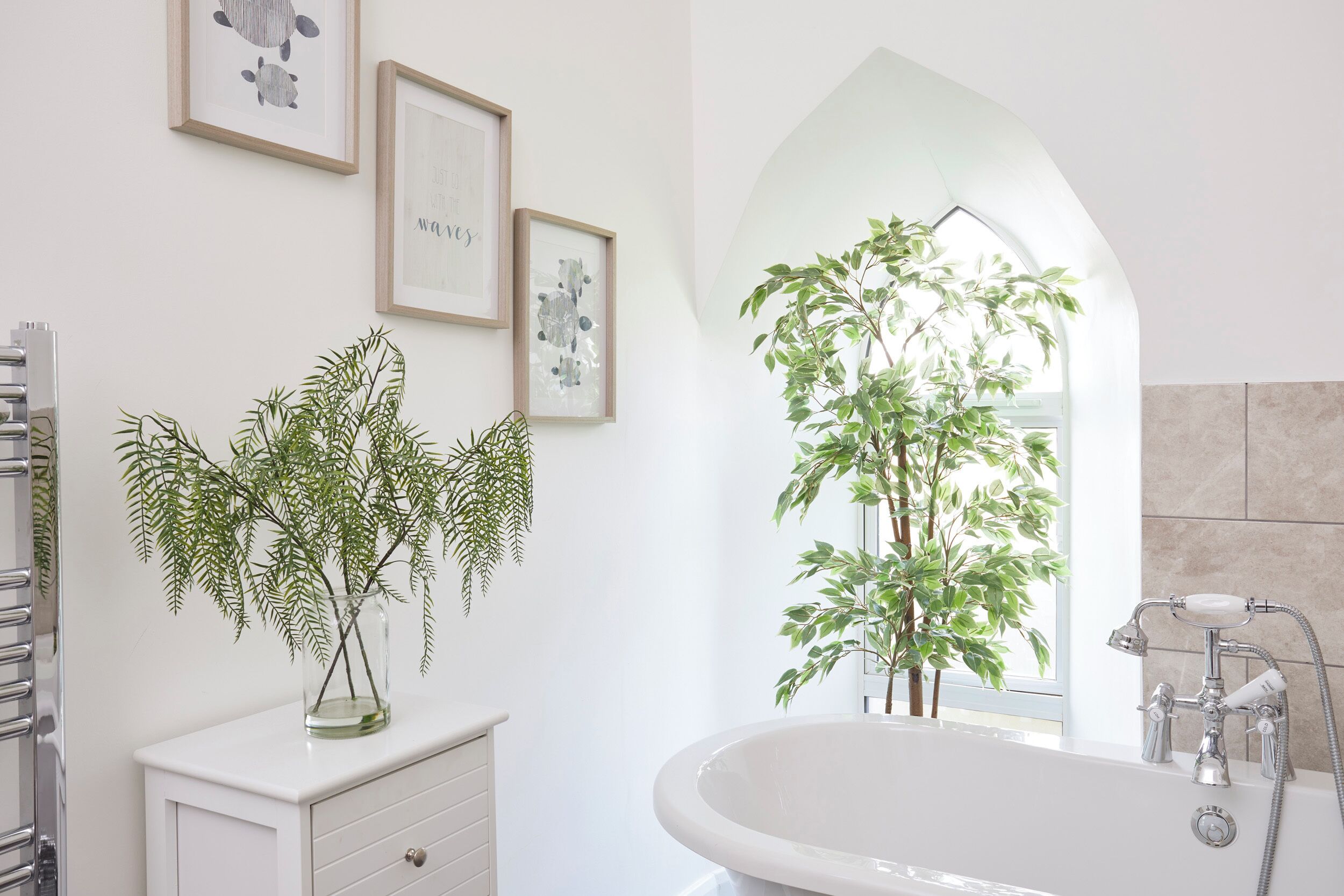 Une salle de bain avec une baignoire, un meuble-lavabo avec des tiroirs, des œuvres d’art encadrées au mur et une plante en pot.