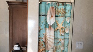 Een badkamer met een douchegordijn met een motief van schelpen en zeesterren, een houten kast, een aanrecht met een waterkoker, en een betegelde vloer.