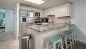Una cocina moderna con armarios blancos, electrodomésticos de acero inoxidable, encimera de granito y taburetes de bar color turquesa.