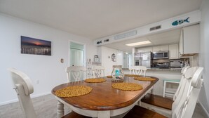 Un comedor con una mesa de madera, sillas blancas y una cocina al fondo.