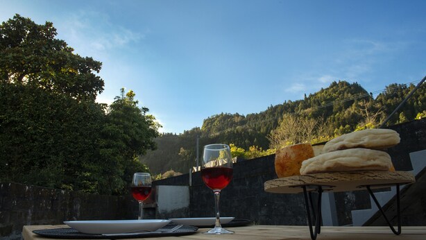 A wooden table with two wine glasses and a loaf of bread, set against a backdrop of a hillside and a clear sky. - Furnas Sweet Home - Central Zone (Furnas)