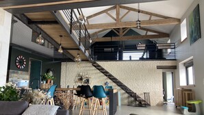 Een ruime loft met een hoge nok, zichtbare houten balken, een entresol en een moderne keukenhoek.