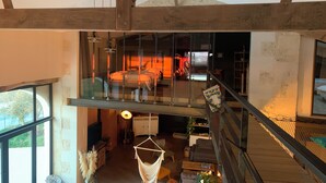 Een ruime loft met een hoge plafon, zichtbare houten balken, een glazen balkon en een gezellige zithoek met kussens en een hangstoel.