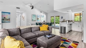 A modern living room with a gray sofa, a coffee table, and a colorful rug. There is a dining area with a table and chairs, and a kitchen with white cabinets and a stainless steel sink.