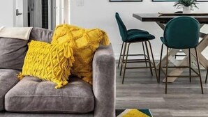 A modern living room with a gray sofa, a yellow throw, and a colorful rug. There is a dining area with a table and chairs, and framed pictures on the wall.