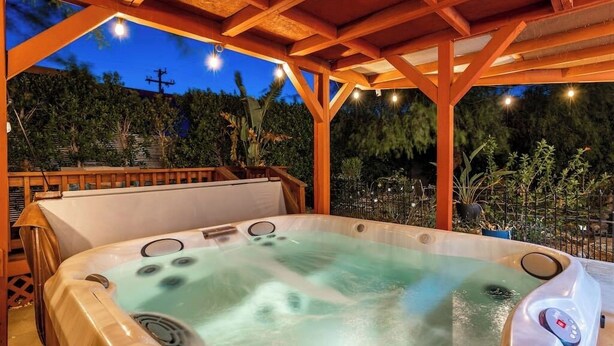 A hot tub under a wooden pergola with a view of a landscaped garden at dusk.
