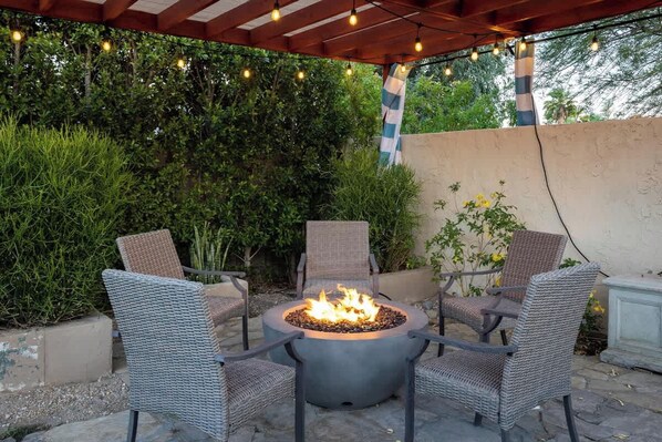 An outdoor seating area with a fire pit surrounded by wicker chairs, under a wooden pergola with string lights.