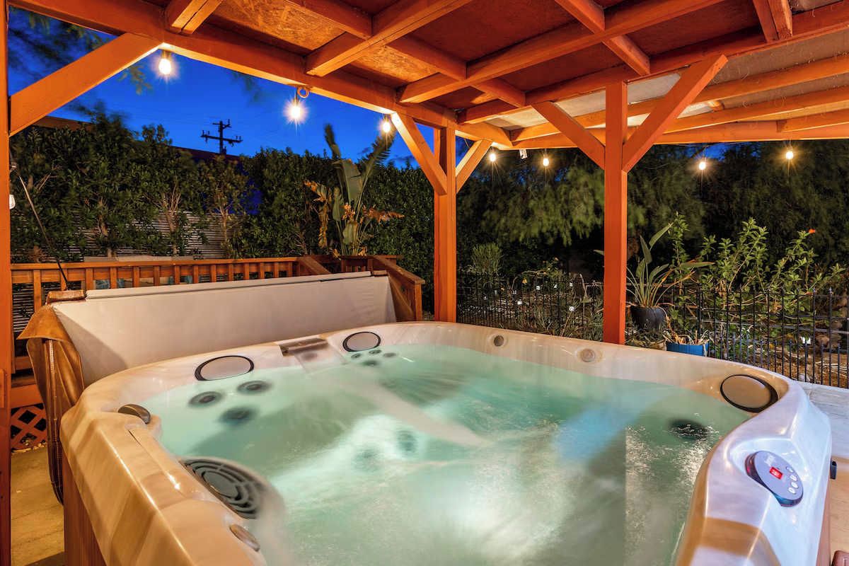 A hot tub under a wooden pergola with a view of trees and a power line in the background.