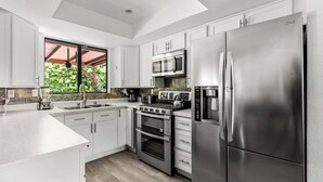 A modern kitchen with white cabinets, stainless steel appliances, and a large window providing a view of greenery.